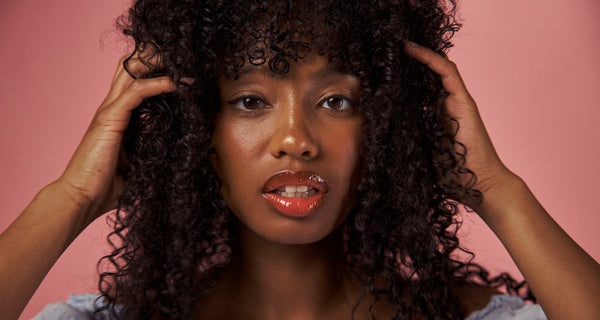 Beauty-Model mit lockigen Haaren und glänzenden, roséfarbenen Lippen vor einem hellrosa Hintergrund.