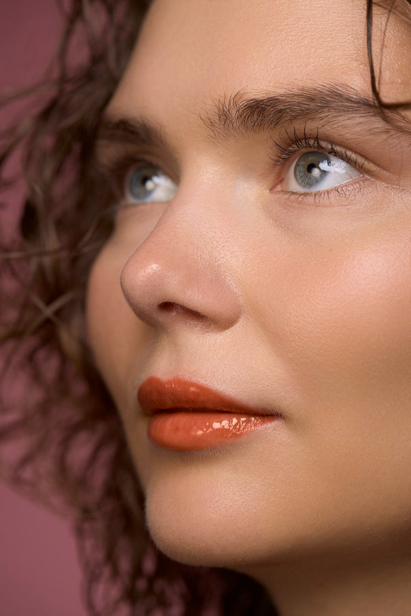 Nahaufnahme eines Beauty-Models mit blauen Augen, lockigen Haaren und glänzenden, orange-korallfarbenen Lippen vor rosa Hintergrund.