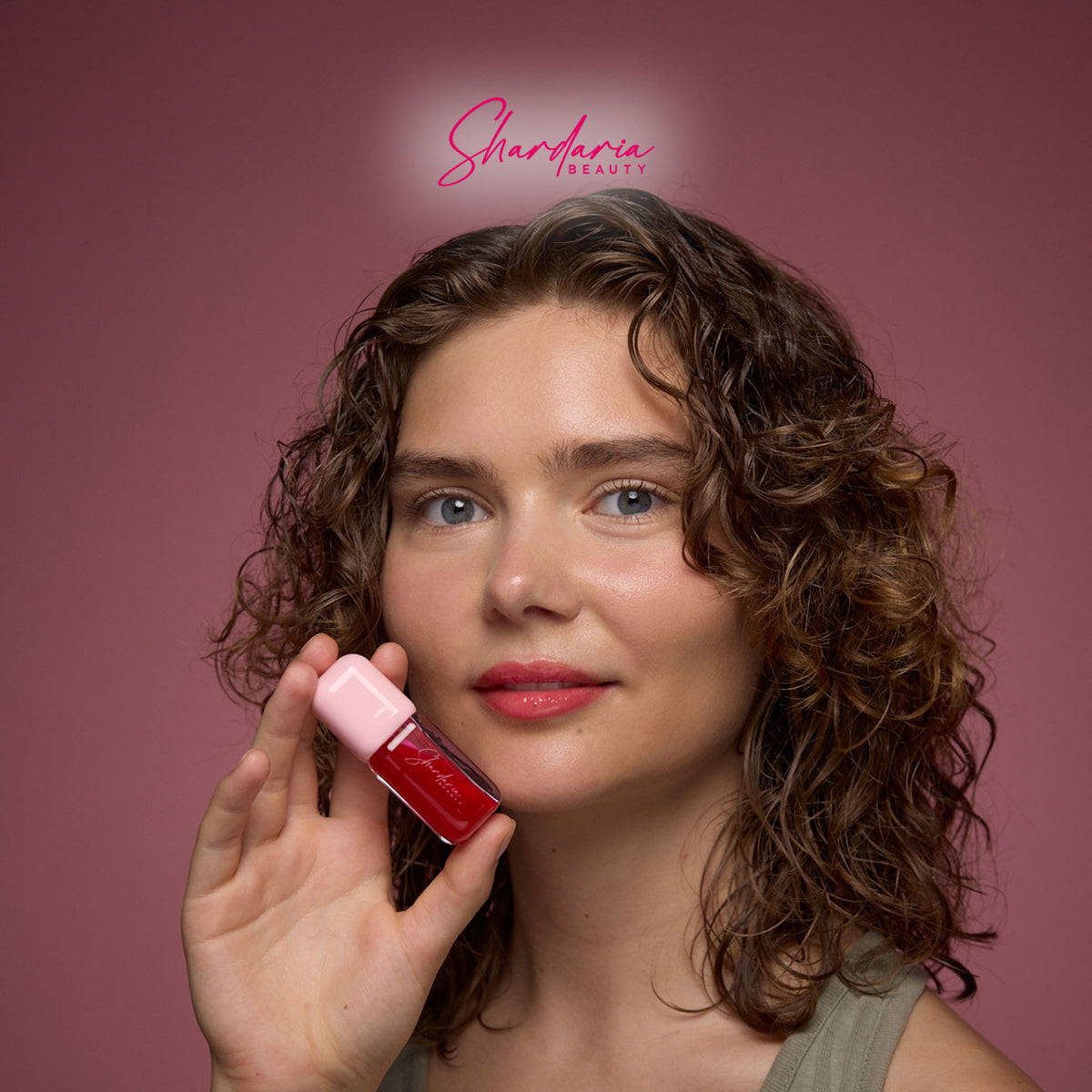 Model hält Cherry Charm Lip Gloss von Shardaria Beauty vor rosafarbenem Hintergrund