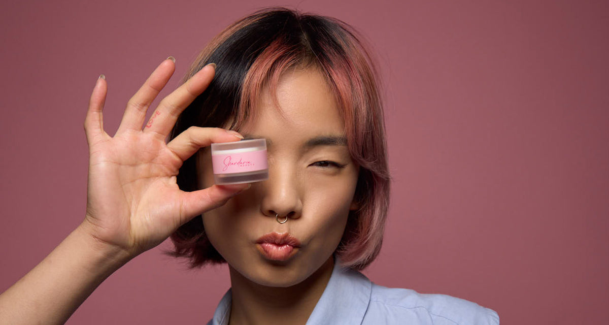 Beauty-Model mit rosafarbenen Haaren hält einen rosa Lippenpflege-Tiegel von Shardaria Beauty vor ihr Auge, vor pinkem Hintergrund.